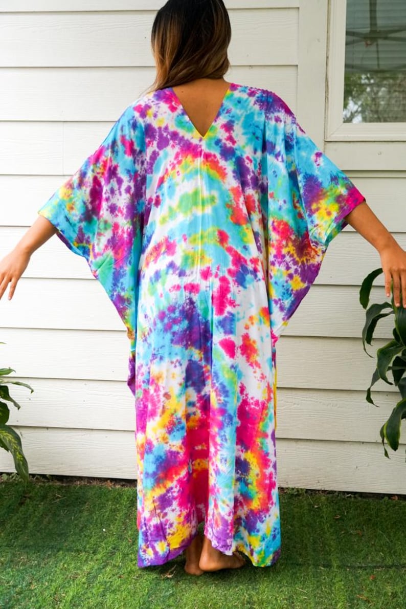 Op de afbeelding: Een enkellange kaftan met een levendig tie-dye patroon in tinten van felroze, elektrisch blauw, zonnig geel en dieppaars. Het kledingstuk heeft een V-hals, wijde mouwen en een losse, ontspannen pasvorm die soepel naar beneden valt.