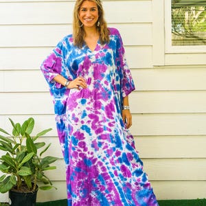 Op de afbeelding: Een enkellange kaftan met een levendig tie-dye patroon in tinten helderblauw, dieppaars en wit. Het kledingstuk heeft een V-hals, wijde mouwen en een losvallend silhouet. De tekst Isla en Amethyst Sky zijn in de afbeelding zichtbaar.