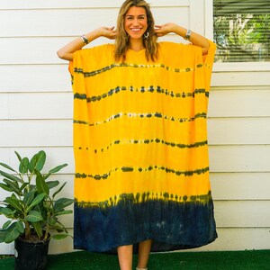 Op de afbeelding: Een felgele kaftan-jurk met horizontale donkerblauwe tie-dye strepen over het lichaam en een effen donkerblauwe zoom aan de onderkant. Het kledingstuk heeft een los, vloeiend silhouet met wijde mouwen en een V-halslijn, gelabeld Isla en Golden Horizon.