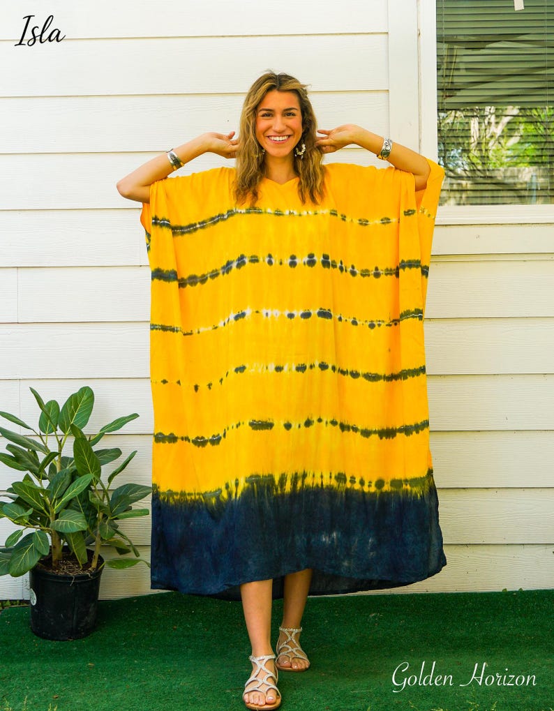 Op de afbeelding: Een felgele kaftan-jurk met horizontale donkerblauwe tie-dye strepen over het lichaam en een effen donkerblauwe zoom aan de onderkant. Het kledingstuk heeft een los, vloeiend silhouet met wijde mouwen en een V-halslijn, gelabeld Isla en Golden Horizon.