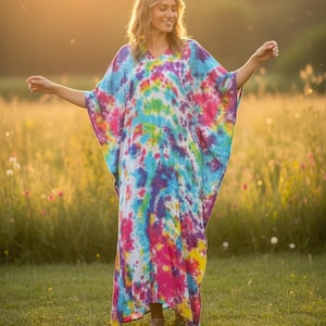 Op de afbeelding: Een enkellange kaftan-jurk met een levendig tie-dye patroon in tinten van felroze, elektrisch blauw, zonnig geel en dieppaars. Het kledingstuk heeft een los, vloeiend silhouet met wijde mouwen en een V-halslijn.
