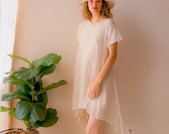 May include: A woman in a cream-coloured, short-sleeved dress with an asymmetrical hemline and a straw hat. The dress has a V-neck and a loose fit. A potted plant and a woven basket are in the background.