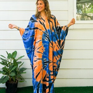 Op de afbeelding: Een vloerlange kaftan met een levendig tie-dye patroon in tinten feloranje, diepblauw en zwart. Het kledingstuk heeft een V-hals, wijd uitlopende mouwen en een losvallend silhouet. De tekst "Isla" en "Blue Horizon" is in de afbeelding aanwezig.