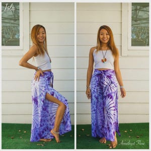 May include: A white sleeveless crop top paired with high-waisted, wide-leg pants featuring a purple and white tie-dye sunburst pattern and side slits. The outfit includes a long necklace with a circular pendant and a silver-toned bracelet.