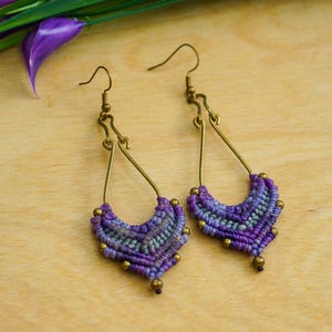 May include: A pair of handmade macrame earrings. The earrings feature a teardrop shape with a purple and blue woven design, accented with small gold-colored beads. The earrings hang from gold-colored ear wires. The background is a light wood surface.
