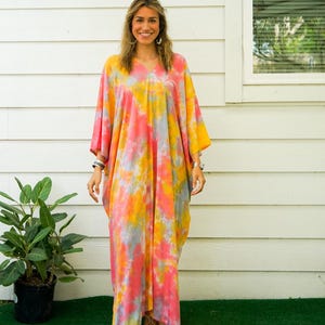 Op de afbeelding: Een enkellange kaftan-jurk met een levendig tie-dye patroon in tinten koraalroze, felgeel en zachtgrijs. Het kledingstuk heeft een V-hals, wijde mouwen en een losvallend silhouet dat tot aan de enkels reikt.