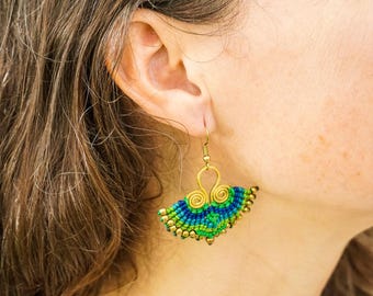May include: A pair of green and blue beaded earrings with a gold wire design. The earrings are hanging from a person's ear.