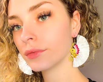 May include: Close-up of a person wearing white, fan-shaped earrings. The earrings have gold wire accents and pink and yellow details. The person has curly blonde hair and is wearing a black top.