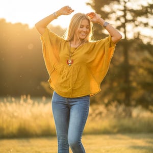 May include: A mustard yellow top with a V-neck and loose sleeves, worn with blue jeans. A pendant necklace with a circular design adds a pop of colour. The outfit is casual and suitable for outdoor wear.