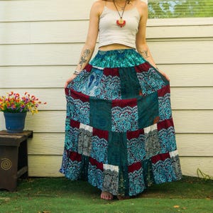 Puede incluir: Una falda larga de patchwork con un estampado de cachemira en tonos turquesa, burdeos y gris. La falda está hecha de múltiples paneles rectangulares y tiene un estilo bohemio fluido. La falda se usa con un top corto blanco.