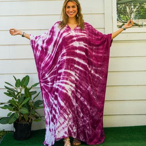 Op de afbeelding: Een vloerlange kaftan met een levendig magenta en wit tie-dye patroon, voorzien van een V-hals en wijde, vloeiende mouwen. Het kledingstuk toont een stralend spiraalontwerp dat zich over de stof uitstrekt voor een opvallend contrast.