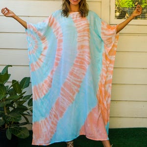 Op de afbeelding: Een losvallende kaftan-jurk met een tie-dye patroon van lichtblauwe en zachte koraaloranje wervelingen. Het kledingstuk heeft wijde, vloeiende mouwen en een ronde halslijn. De tekst 'Isla' en 'Coral Reef' staat in de hoeken van de afbeelding.
