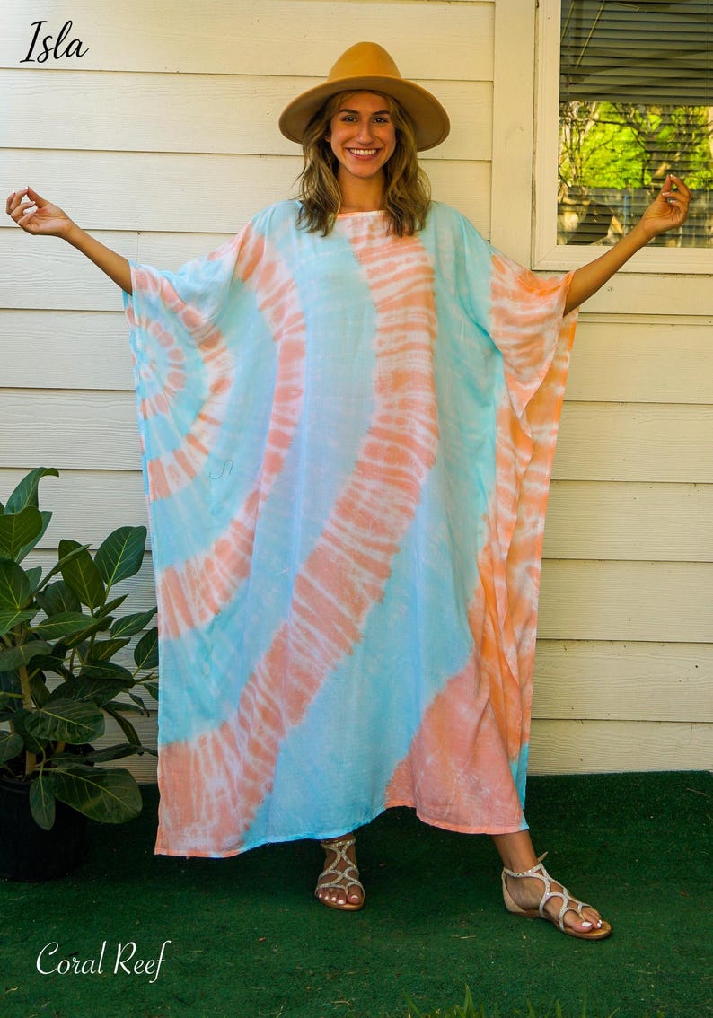 Op de afbeelding: Een losvallende kaftan-jurk met een tie-dye patroon van lichtblauwe en zachte koraaloranje wervelingen. Het kledingstuk heeft wijde, vloeiende mouwen en een ronde halslijn. De tekst 'Isla' en 'Coral Reef' staat in de hoeken van de afbeelding.