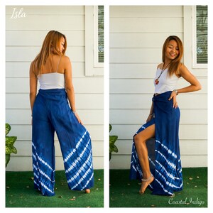 May include: A pair of wide-leg pants in a deep indigo blue featuring a white tie-dye diagonal stripe pattern. The pants have a high-waisted design with a front tie closure and a high side slit that reveals the leg. The fabric is lightweight and flowy.