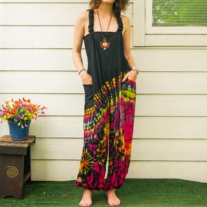 May include: A woman wearing a black tie-dye jumpsuit with a colorful swirl pattern. The jumpsuit has a wide leg and a relaxed fit. She is standing in front of a white wall with a window behind her.
