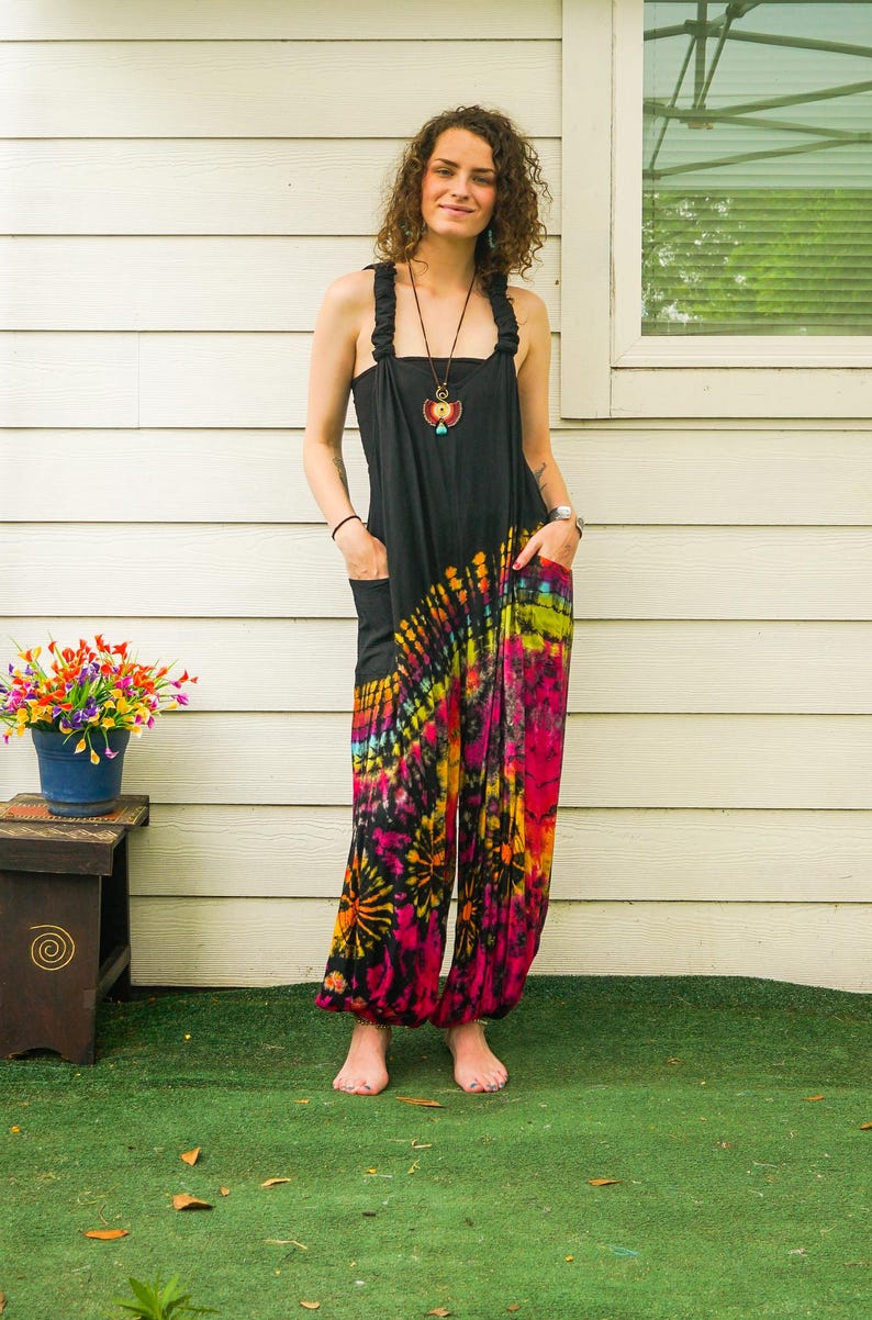 May include: A woman wearing a black tie-dye jumpsuit with a colorful swirl pattern. The jumpsuit has a wide leg and a relaxed fit. She is standing in front of a white wall with a window behind her.