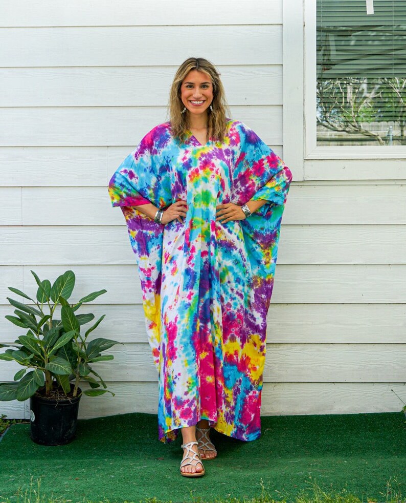 Op de afbeelding: Een enkellange kaftan-jurk met een levendig tie-dye patroon in tinten felroze, elektrisch blauw, zonnig geel en wit. Het kledingstuk heeft een los, vloeiend silhouet met wijde mouwen, een V-hals en een ontspannen, comfortabele pasvorm.