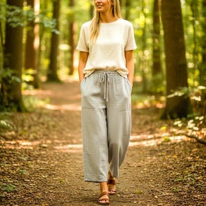 May include: A short-sleeved, cream-colored linen top paired with loose-fitting, light gray linen trousers featuring a drawstring waist, two front pockets, and a wide-leg cut, complemented by brown leather sandals with two horizontal straps.