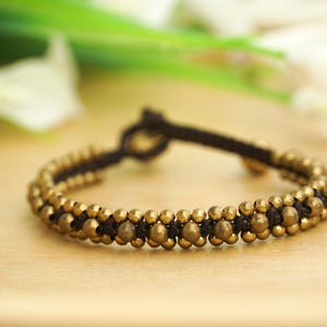 May include: A bracelet featuring small, round, gold-coloured beads strung on a black cord. The bracelet is knotted at the ends and is displayed on a light-coloured surface. The background is blurred, with hints of green and white.