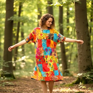May include: A colorful patchwork dress with short sleeves and a flared hem. The dress features a vibrant mix of red, blue, yellow, and orange fabric squares. The person is wearing brown sandals and walking in a forest.