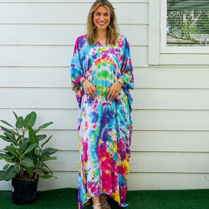 Op de afbeelding: Een enkellange kaftan-jurk met een levendig tie-dye patroon in tinten van felroze, elektrisch blauw, zonnig geel en paars. Het kledingstuk heeft een V-hals, losvallende korte mouwen en een ontspannen, vloeiend silhouet.