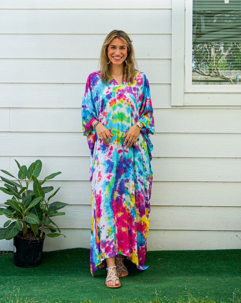 Op de afbeelding: Een enkellange kaftan-jurk met een levendig tie-dye patroon in tinten van felroze, elektrisch blauw, zonnig geel en paars. Het kledingstuk heeft een V-hals, losvallende korte mouwen en een ontspannen, vloeiend silhouet.