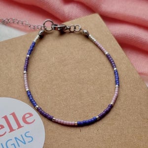 Op de afbeelding: Een delicate kralenarmband met een zilveren sluiting, met een patroon van blauwe, paarse, roze en zilveren kralen. De armband ligt op een bruin kartonnen oppervlak met een witte sticker met de tekst "giselle DESIGNS". Een roze stof is op de achtergrond.
