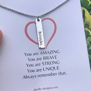 May include: A silver-colored necklace with a rectangular pendant that reads "breathe" in a vertical orientation. The pendant hangs from a delicate chain. The necklace is displayed on a white card with a pink heart and inspirational text.