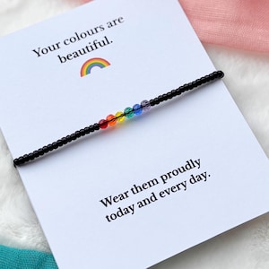 May include: A black beaded bracelet with rainbow-coloured beads rests on a white card. The card features the text "Your colours are beautiful" and "Wear them proudly today and every day." A rainbow graphic is also present.