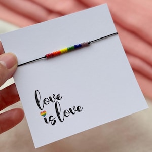 Peut inclure: Un bracelet artisanal avec des perles arc-en-ciel sur un cordon noir, présenté sur une carte blanche. La carte porte l'inscription "love is love" avec un cœur arc-en-ciel. Le fond est un tissu rose.