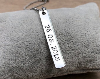 May include: A silver bar pendant necklace with the date "26.08.2018" engraved vertically. The pendant hangs on a delicate chain and rests on a gray, plush surface.
