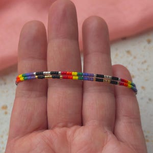 Pode incluir: Duas pulseiras de contas, cada uma com um padrão de cores do arco-íris, são exibidas nos dedos. As pulseiras apresentam pequenas contas coloridas em uma sequência repetida de vermelho, laranja, amarelo, verde, azul e roxo, separadas por contas pretas e prateadas. Um acessório vibrante.
