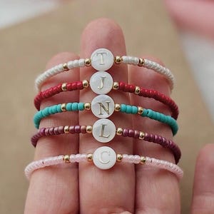 Può includere: Sono presentati sei braccialetti di perline in bianco, rosso, turchese, bordeaux e rosa. Ogni braccialetto presenta un ciondolo circolare bianco con una lettera. Perline color oro accentuano i braccialetti.