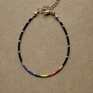 Könnte beinhalten: Ein zartes Armband mit einem schwarzen Perlenband, goldenen Akzenten und einem Regenbogenfarbenen Abschnitt. Das Armband hat eine goldfarbene Kette zur Größenanpassung und einen Karabinerverschluss. Das Armband wird auf einer neutralen Oberfläche präsentiert.