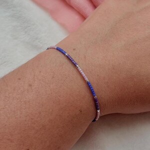 May include: A slender beaded bracelet in blue, pink, and silver. The bracelet features small, evenly spaced beads, forming a minimalist and elegant design. The bracelet is worn on a wrist.