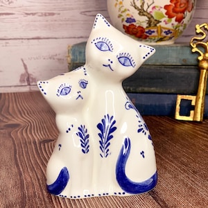 May include: A white ceramic figurine of two cats, one larger than the other, with blue and white painted details. The cats have blue eyes and are sitting side-by-side.