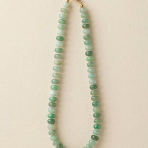 May include: A necklace featuring a single strand of smooth, round, light and dark green beads. The beads are strung together and secured with a gold-toned metal lobster claw clasp at the top, creating a circular shape for the jewelry piece.