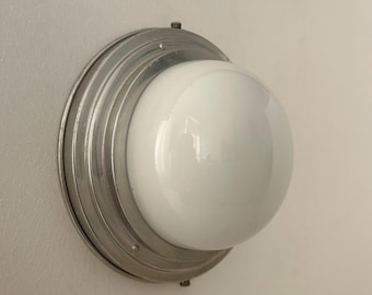 May include: A vintage-style wall sconce featuring a large, spherical white glass shade. The shade is set within a circular, ridged metal fixture. The fixture is mounted on a white wall, creating a classic lighting design.