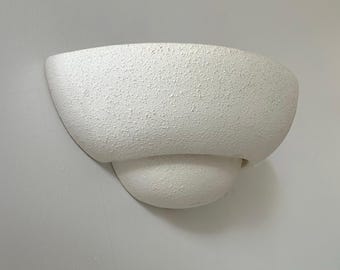 Puede incluir: Aplique de pared blanco texturizado, con una parte superior redondeada en forma de cuenco y una sección inferior esférica más pequeña. El aplique tiene una superficie rugosa y moteada, montado en una pared blanca. Diseño moderno y minimalista.