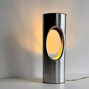 May include: A silver cylindrical table lamp with a large oval cutout in the center. The lamp is lit and casting a warm glow.