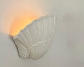 May include: A white wall-mounted sconce shaped like a seashell with a textured, ribbed surface. The fixture emits a warm, soft orange glow from its interior, casting light upward and outward against the surrounding wall surface.