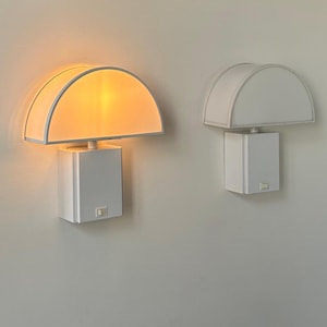 May include: Two white wall sconces with a semi-circular shade and a rectangular base. One sconce is illuminated with a warm light, while the other is unlit. The shades are white with a white trim, and the base is a white square.