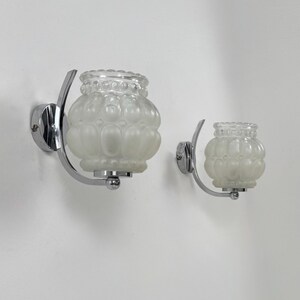 May include: Two vintage wall sconces with frosted glass shades and chrome-finished metal hardware. The glass shades have a textured, bubble-like design. The sconces are mounted on a white wall.