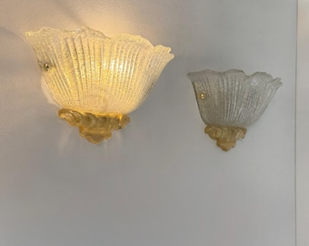 May include: Two wall sconces with a textured, petal-shaped glass shade and a gold-colored base. The sconces are mounted on a white wall. The larger sconce is lit, emitting a warm glow. The smaller sconce is unlit.