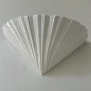 Puede incluir: Aplique de pared blanco en forma de abanico, con una superficie texturizada y acanalada. El aplique está hecho de cerámica o porcelana y está montado en una pared blanca. El diseño se asemeja a una concha.