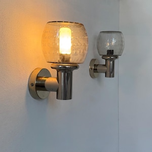May include: Two wall-mounted sconces. One is lit, with a warm glow emanating from a bulb inside a textured, amber-colored glass globe. The other sconce has a gray glass globe. Both have a brushed metal base and are mounted on a white wall.