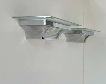 May include: Two wall-mounted shelves featuring clear glass tops and polished chrome metal brackets. Each bracket has a curved, ribbed design that supports the rectangular glass surface, creating a sleek, reflective appearance for wall storage.