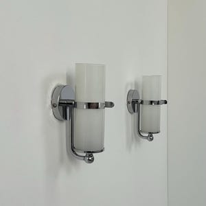 May include: Two wall sconces with a chrome finish and cylindrical frosted glass shades. The sconces are mounted on a white wall. The design is minimalist and modern, suitable for interior lighting.