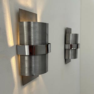 Könnte beinhalten: Zwei Wandleuchten aus gebürstetem Metall. Jede Leuchte hat einen zylindrischen Körper mit einem polierten Metallband und einer rechteckigen Rückplatte. Die Leuchten sind an einer weißen Wand montiert.
