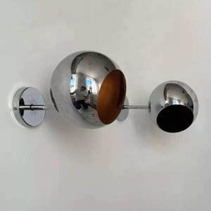 Peut inclure: Une applique murale chromée avec deux abat-jours sphériques. Le plus grand abat-jour a une ouverture découpée, tandis que le plus petit abat-jour est plein.